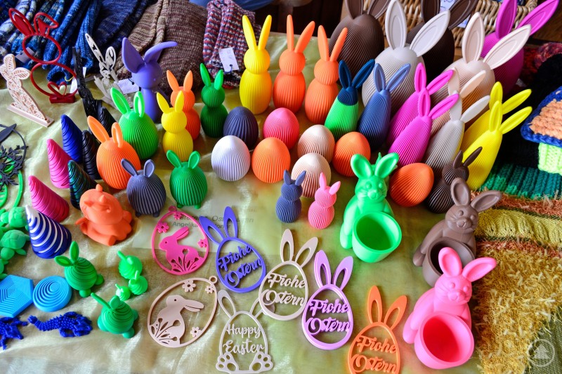 Osterhasen aus dem 3D Drucker Das Bild zeigt einen farbenfrohen Ostermarkt-Stand beim Eiermarkt 2025, auf dem eine Vielzahl an dekorativen Hasenfiguren, Ostereiern und Anhängern aus Kunststoff oder 3D-Druckmaterialien angeboten wird. Die Figuren sind in kräftigen Farben wie Orange, Pink, Grün, Blau oder Lila gestaltet, teils geriffelt, teils glatt, mit aufgesetzten Ohren oder eingravierten Ostergrüßen wie „Frohe Ostern“ oder „Happy Easter“. Dazwischen liegen gestrickte Waren und filigrane Holzanhänger. Die bunte Vielfalt und Kreativität der Stücke machen den Stand zu einem echten Hingucker – modern, verspielt und dennoch thematisch ganz im Zeichen des Osterfestes.