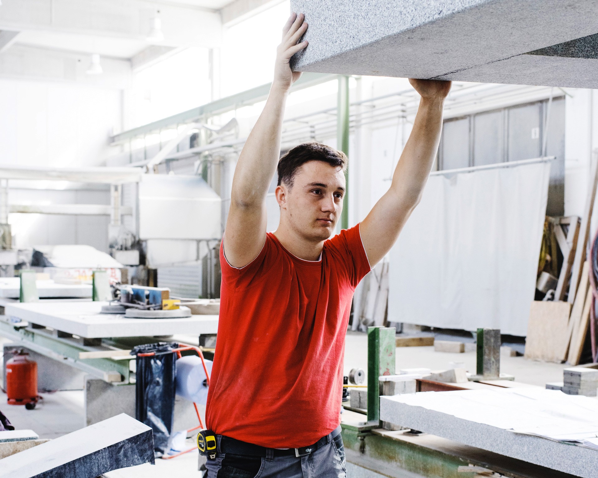 Das Bild zeigt einen jungen Mann in einem industriellen Arbeitsumfeld, vermutlich in einer Werkstatt oder Fertigungshalle. Er trägt ein rotes T-Shirt und hält eine große Steinplatte oder ein schweres Material über seinem Kopf. Im Hintergrund sind Maschinen und Arbeitsmaterialien zu sehen, was auf eine Produktionsumgebung hinweist. Das Bild vermittelt einen Eindruck von handwerklicher Arbeit und körperlicher Anstrengung.