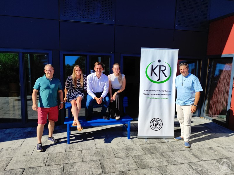 Diskutierten u. a. aktuelle gesellschaftliche und politische Entwicklungen und Veränderungen: (v. li. n. re.) Stellv. KJR-Vorsitzender Armin Wildfeuer, KJR-Geschäftsführerin Michaela Pertler, MdL Dr. Stefan Ebner, KJR-Vorstandsmitglied Alina Stockinger und KJR-Vorstandsmitglied Tim Weidinger. Fünf Personen stehen bzw. sitzen neben einem Roll-Up des Kreisjugendrings vor einem modernen Gebäude in der Sonne und blicken in die Kamera.