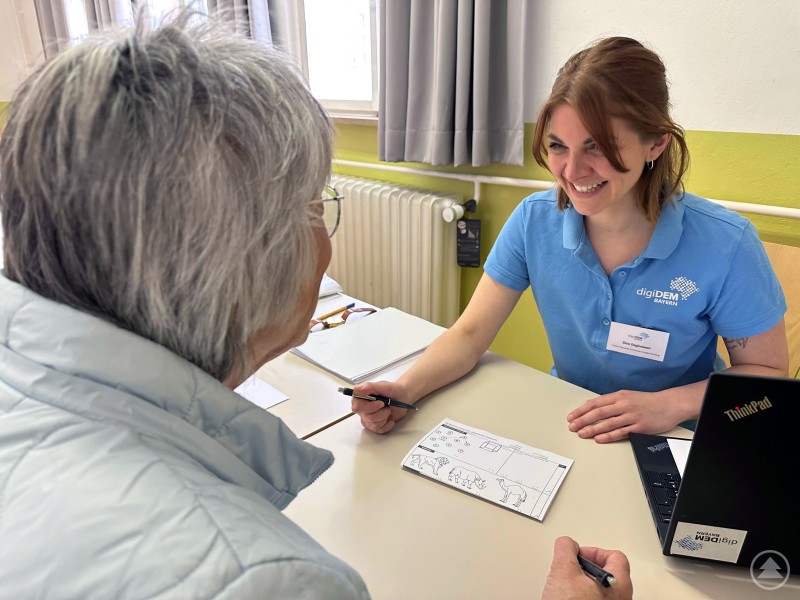 Der Demenztest wird von geschulten Mitarbeitenden durchgeführt und liefert erste Hinweise auf eine mögliche weitere ärztliche Abklärung. Eine Mitarbeiterin von digiDEM Bayern sitzt an einem Tisch, lacht freundlich und erklärt einer älteren Frau einen Gedächtnistest. Vor ihr liegen Unterlagen und ein Laptop.