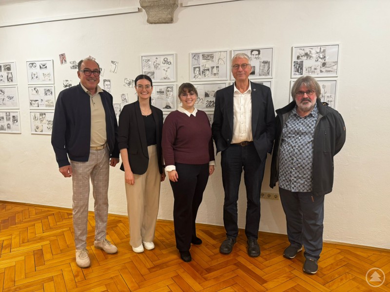 Stellvertretender Landrat Franz Brunner, Musikerin Antonia Höpfl, Kulturreferentin Gabriela Rauscher, Künstler Helmut Wartner und Laudator Hans Haslsteiner in der Sonderausstellung „Jeden Tag eine Postkarte“. Fünf Personen stehen in einer Ausstellung vor einer Wand mit gezeichneten Postkarten. Sie blicken in die Kamera und lächeln. Der Raum hat einen Holzboden, und an der Wand hängen mehrere gerahmte Zeichnungen.