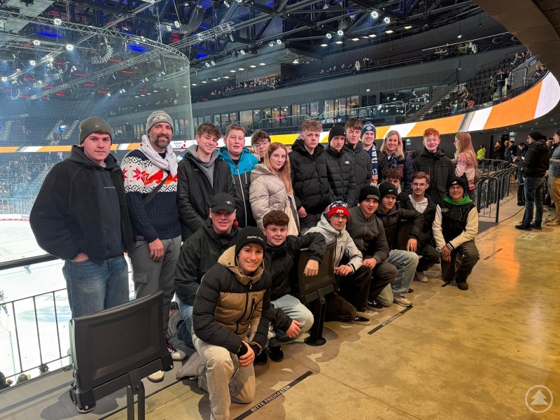 Ein besonderes Highlight war für die Realschüler auch das Eishockeyspiel des EHC Red Bull München in der SAP-Arena gegen die Kölner Haie. Eine Gruppe Jugendlicher und Erwachsener posiert am Rand eines Eishockeystadions. Im Hintergrund sind Eisfläche, Bande und Zuschauerränge des SAP Garden zu erkennen.