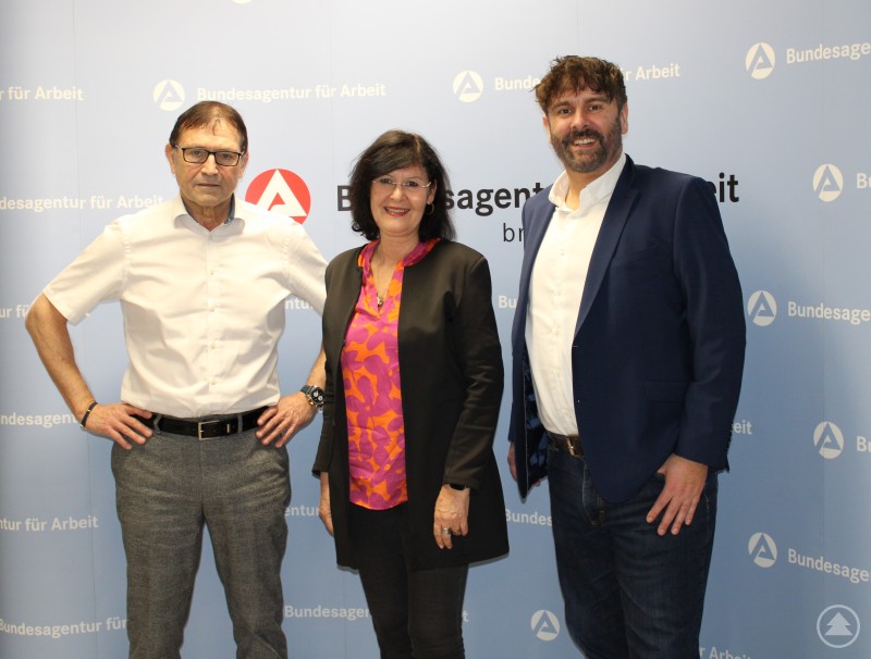 Von links: Hermann Käser, Eva-Maria Kelch, Tobias Cziudaj Drei Personen stehen vor einer blauen Wand mit dem Logo der Bundesagentur für Arbeit. Links ein Mann im weißen Hemd, in der Mitte eine Frau in dunkler Jacke und farbigem Oberteil, rechts ein Mann im dunklen Sakko. Alle blicken in die Kamera und posieren für ein offizielles Foto.