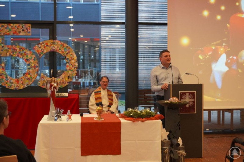 Adventsgottesdienst der Wolfsteiner Werkstätten: Feierlicher Rückblick und 50-jähriges Jubiläum im Zeichen von Licht und Hoffnung. Ein Mann steht an einem Rednerpult mit Caritas-Logo und spricht vor Publikum, neben ihm sitzt ein Geistlicher im liturgischen Gewand an einem festlich gedeckten Tisch mit Adventskranz und Kerzen. Im Hintergrund sind eine große bunte "50" als Jubiläumszahl sowie weihnachtliche Dekorationen und eine Projektionsfläche mit Kerzenmotiv zu sehen. Der Raum ist modern mit großen Fenstern.