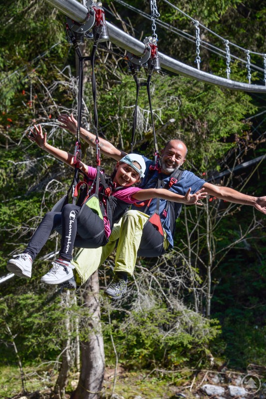 Flyline am Almberg: Mit der geplanten Fertigstellung und Inbetriebnahme bis zum Sommer wird das Angebot im Skizentrum Mitterdorf künftig auch auf den Sommerbetrieb ausgeweitet. Ein Mann und ein Kind gleiten gesichert an einer Flyline durch den Wald und genießen sichtlich die Fahrt.