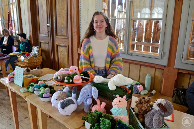 Maria Obermüller jüngste Teilnehmerin Das Bild zeigt eine junge Frau mit einem bunten Strick-Cardigan, die an ihrem liebevoll gestalteten Verkaufsstand beim Eiermarkt 2025 sitzt. Vor ihr sind zahlreiche selbstgehäkelte Kuscheltiere in fröhlichen Farben ausgestellt – darunter Hasen, Katzen, Dinosaurier, Vögel, Schnecken und Frösche. Die Figuren bestehen aus weichem Chenille-Garn und wirken einladend und detailreich gearbeitet. Im Hintergrund sind weitere Marktbesucher zu sehen, darunter ein Junge mit Korb sowie eine ältere Frau im Gespräch. Die Szene spiegelt Kreativität, Handarbeit und ein generationenübergreifendes Markterlebnis in gemütlicher Atmosphäre wider.