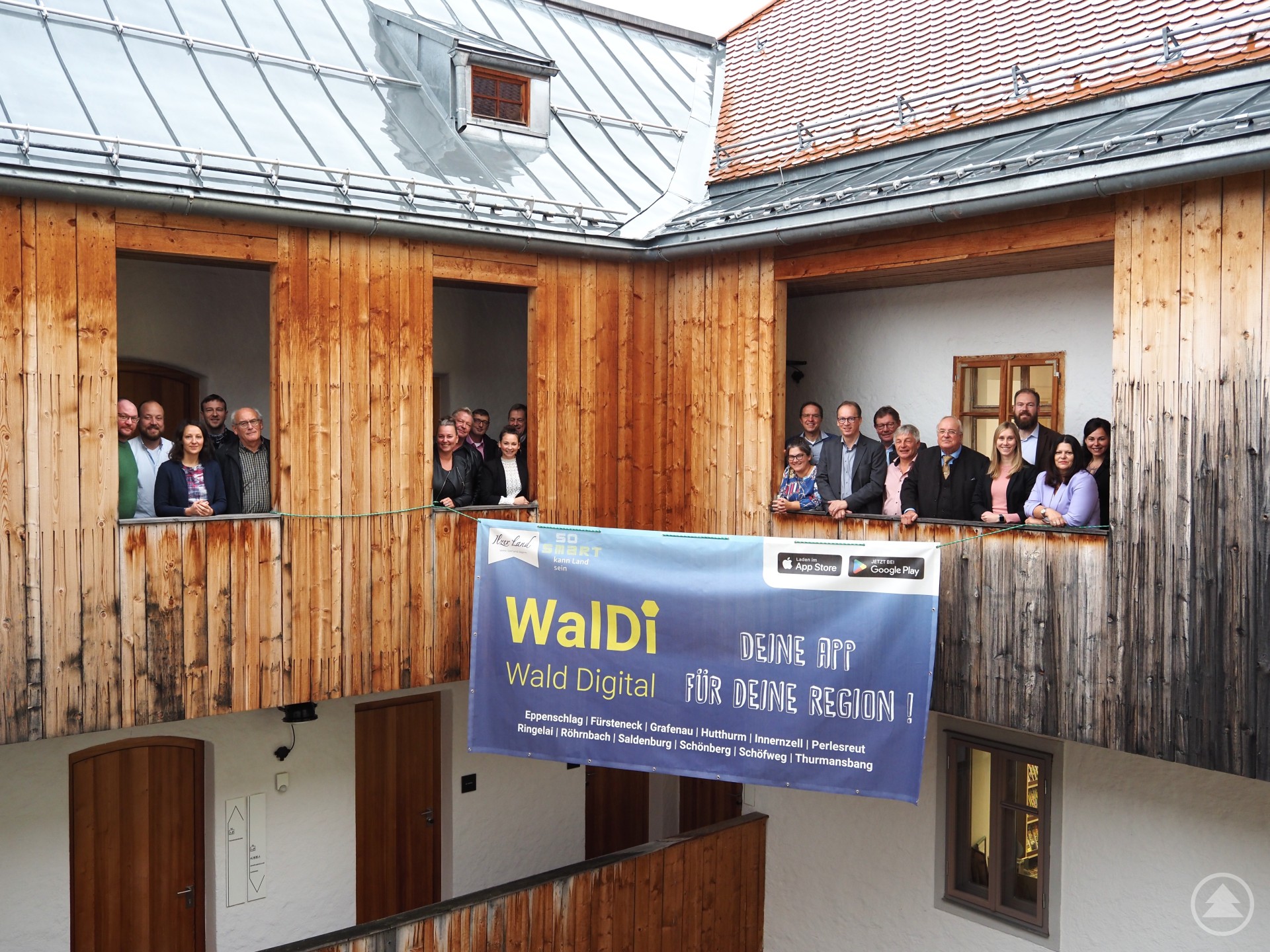 Gruppenbild der ILE Ilzer Land in der Bauhütte Perlesreut, der Geschäftsstelle der ILE. Eine Gruppe von Menschen steht auf einem Balkon in einem Gebäude mit Holzfassade und einem großen Banner mit der Aufschrift "WalDi – Wald Digital".