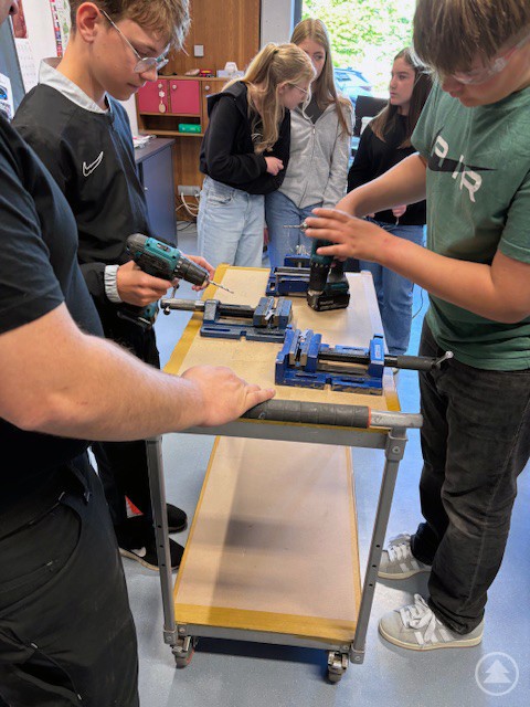 Schüler hantieren unter Anleitung mit Bohrmaschinen und Schraubstöcken an einem Werkstattwagen.