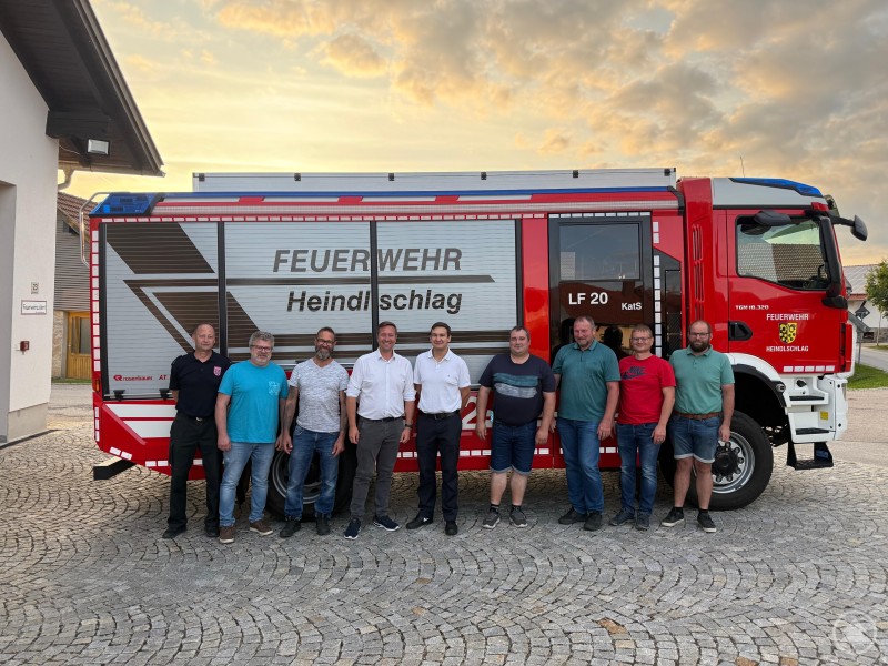 Kreisbrandmeister Martin Bauer, Alfred Seibold, Ludwig Schätzl, MdL Josef Heisl, Maximilian Meier, Roland Parockinger, Bürgermeister Roland Freund, Florian Scharinger und Franz Hauer (v.l.) vor dem Feuerwehrfahrzeug der FFW Heindlschlag Neun Männer stehen in einer Reihe vor einem modernen roten Löschfahrzeug der Feuerwehr Heindlschlag, das auf einem gepflasterten Platz geparkt ist.