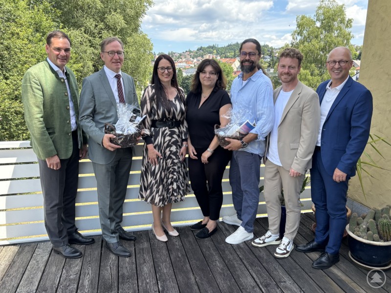 Sebastian Gruber, Landrat von Freyung-Grafenau (v. l.), Michael Hinterdobler, Generalsekretär Wirtschaftsbeirat Bayern, Jaroslava Pongratz, Netzwerkmanagerin Bayern – Böhmen, Manuela Königbauer, Regierung von Niederbayern, Tomáš Studeník, Keynote Speaker, Johannes Gastinger, Wirtschaftsförderer LK Freyung-Grafenau, Kaspar Sammer, Geschäftsführer Euregio und Geschäftsführer Europaregion Donau-Moldau Niederbayern. Sieben Personen stehen auf einer Terrasse mit Blick auf eine Stadt im Hintergrund; zwei von ihnen halten Präsentkörbe in der Hand.