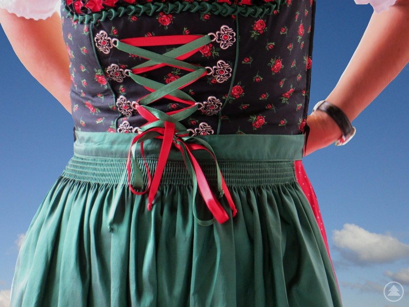 Zu sehen ist das Detail einer traditionellen Tracht: Ein Dirndl mit grüner Schürze und bunt gemustertem Mieder, dessen rote und grüne Bänder kunstvoll zur Schleife gebunden sind. Die Hände der Person ruhen an der Hüfte, im Hintergrund leuchtet ein klarer Himmel. Das Bild vermittelt bayerisches Lebensgefühl und verbindet Tradition mit Individualität.