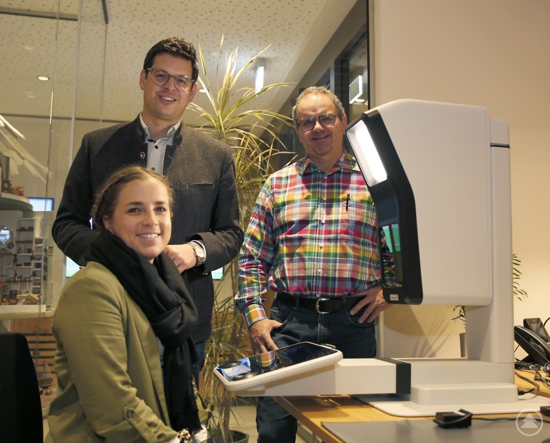 Sophia Bumberger (v.l.), Bürgermeister Martin Pichler und Robert Maier als EDV-Spezialist am Markt durften beim Fototermin auch lächeln – hier ging es um die Vorstellung der neuen „Kollegen“ im Bürgeramt für mehr Service und Vereinfachung von Behördengängen. Drei Personen stehen und sitzen an einem modernen Passbildautomaten im Bürgerbüro. Eine Frau sitzt am Gerät, während zwei Männer danebenstehen und in die Kamera lächeln. Im Hintergrund sind Glaswände, Pflanzen und Büroraum erkennbar.