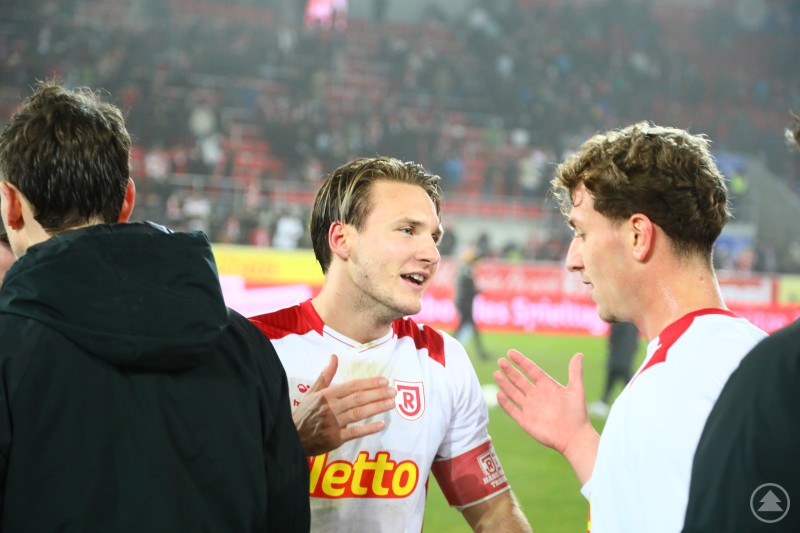 Zwei Spieler des SSV Jahn Regensburg unterhalten sich nach dem Abpfiff auf dem Spielfeld. Beide wirken zufrieden und gestikulieren während des Gesprächs. Im Hintergrund sind leere Ränge und einige Zuschauer zu sehen.