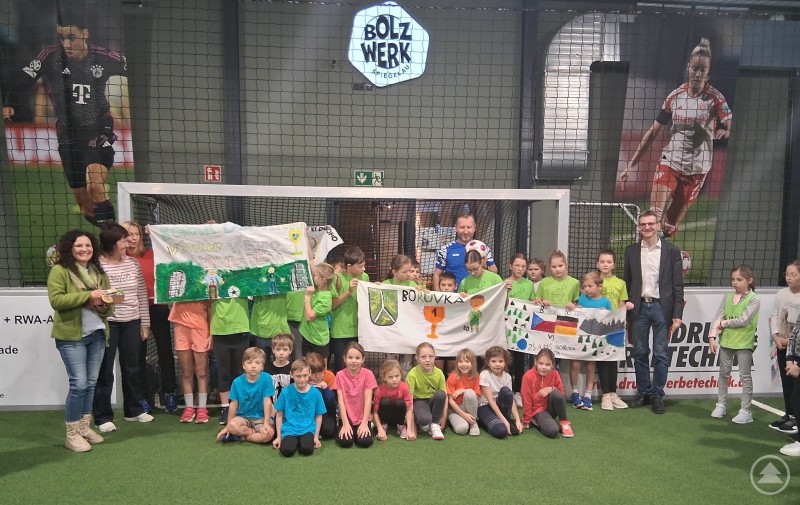 Bürgermeister Karlheinz Roth, Bolzwerkinhaber Michael Miedl und Schulleiterin Corinna Lerach-Wirsich begrüßen die tschechischen Gäste mit Schulleiterin Alena Braunova. Eine große Gruppe von Kindern und Erwachsenen steht in grünen und blauen Shirts in einer Indoor-Fußballhalle vor einem Tor. Einige Kinder halten selbstgemalte Banner mit Motiven zu Sport und deutsch-tschechischer Freundschaft. Im Hintergrund ist das Logo „Bolzwerk Spiegelau“ zu sehen.