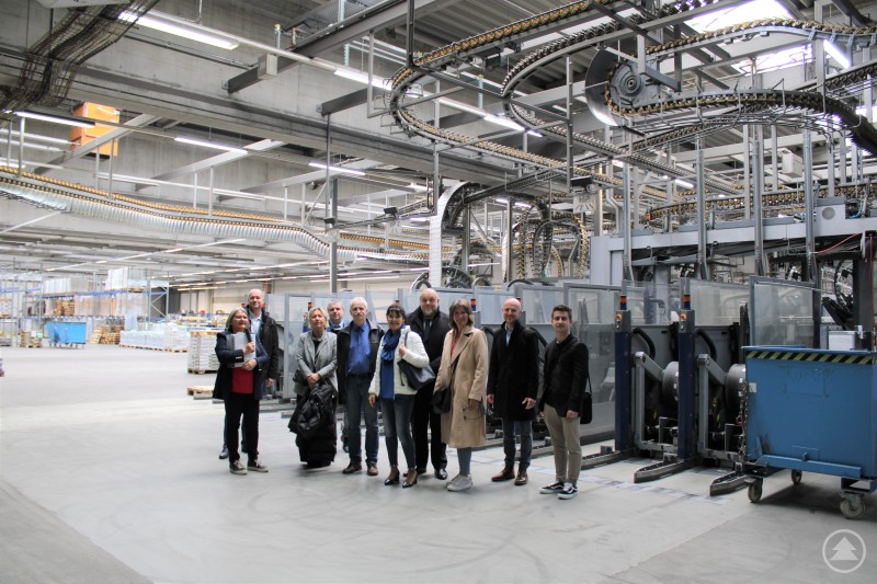 von links: Roswitha Fraunhofer, Jiri Beran, Pavla Blahová, Libor Simanek, Ivan Loukota, Eva-Maria Kelch, Václav Pauch, Christine Reitberger, Gabriel Fredl, Thomas Wirringer Das Bild zeigt eine Gruppe von Besucherinnen und Besuchern bei einer Führung durch ein modernes Medien- oder Logistikzentrum. Sie stehen in einer weitläufigen Halle mit hohem, industriellem Deckengebälk und einer komplexen Förder- und Sortieranlage, die über ihren Köpfen verläuft. Paletten, Rollcontainer und Maschinen sind im Hintergrund sichtbar. Die Gruppe wirkt interessiert und freundlich – es handelt sich vermutlich um eine Delegation aus Verwaltung, Bildung oder Wirtschaft, die sich über Abläufe und Technik vor Ort informiert. Die Szenerie vermittelt Fortschritt, Digitalisierung und effiziente Logistikprozesse in einer hochorganisierten Umgebung.