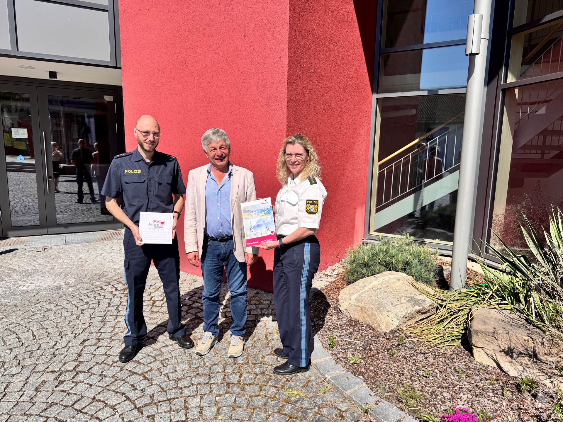 Polizeioberrätin Ingrid Grötzinger, Bürgermeister Leo Meier und Polizeihauptkommissar Andreas Schrank bei der Vorstellung des Sicherheitsberichts in Röhrnbach. Drei Personen stehen vor einem roten Gebäude und halten Broschüren in den Händen. Es handelt sich um zwei Polizeibeamte in Uniform und den Bürgermeister in Freizeitkleidung. Die Szene spielt sich im Sonnenschein auf einem gepflasterten Platz ab.