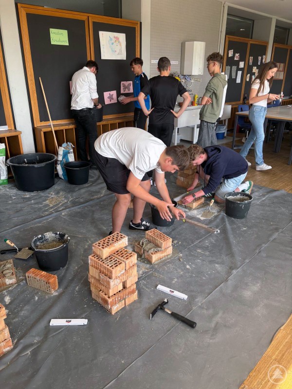 Jugendliche arbeiten mit Mörtel und Ziegelsteinen an kleinen Mauerwerken in einem Klassenraum.