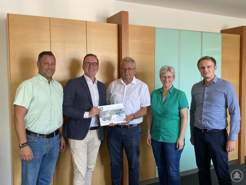 Wirtschaftsförderer Martin Köck, Dr. Reinhard Saller, Landrat Josef Laumer, geschäftsleitende Beamtin Silke Raml und Abteilungsleiter Andreas Käser (v. l.) beim Vorstellungsbesuch im Landratsamt Straubing-Bogen. Fünf Personen stehen in einem Büro vor einer Holz-Glas-Schrankwand, zwei von ihnen halten gemeinsam ein Dokument mit einer Jubiläumsüberschrift.