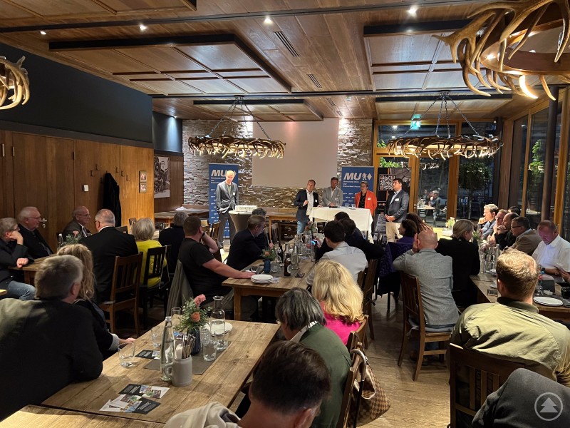 Sommerempfang der Mittelstands-Union mit Impulsvortrag von Prof. Dr. Volker Stockinger. Eine größere Runde von Gästen sitzt in einem Restaurant mit Holzdekor und hört aufmerksam einem Redner auf der Bühne zu. Im Hintergrund stehen Banner der Mittelstands-Union.