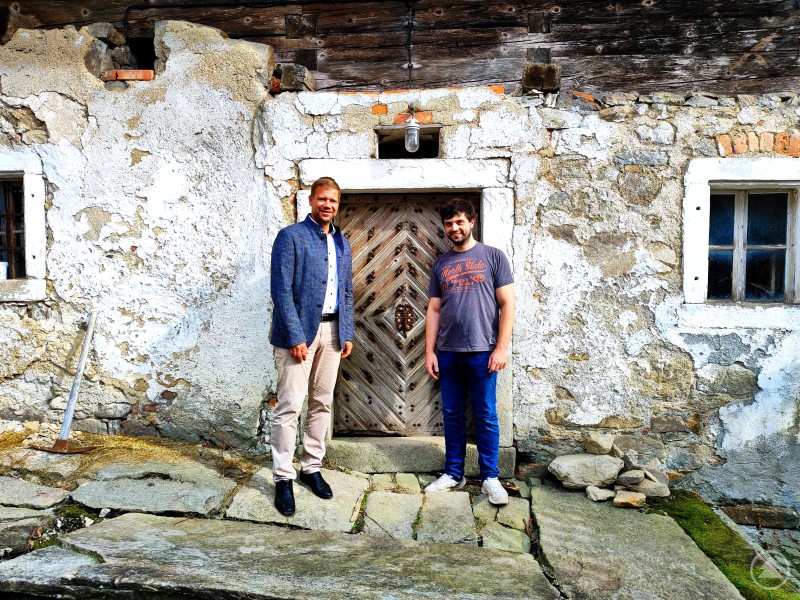 Kreisheimatpfleger Dr. Leonhard Bürger (re.) informiert Bezirkstagspräsident Dr. Olaf Heinrich (li.) über die denkmalgerechte Instandsetzung seines Anwesens in Appmannsberg. Zwei Männer stehen vor einem alten, denkmalgeschützten Bauernhaus mit verwitterter Stein- und Holzfassade. Die Eingangstür aus Holz zeigt ein Fischgrätmuster. Einer der Männer trägt ein Sakko, der andere ein T-Shirt und Jeans. Die Szene vermittelt das Gefühl von Geschichte und Erhalt alter Bausubstanz.