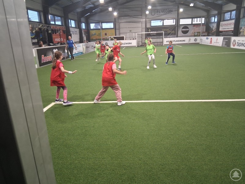 In bayerisch-tschechischen Teams wetteiferten die Schüler gemeinsam um Tore. Mehrere Kinder in roten und grünen Trikots spielen gemeinsam Fußball auf einem Kunstrasenplatz in einer Indoor-Halle. Im Hintergrund beobachtet ein Erwachsener das Spiel.