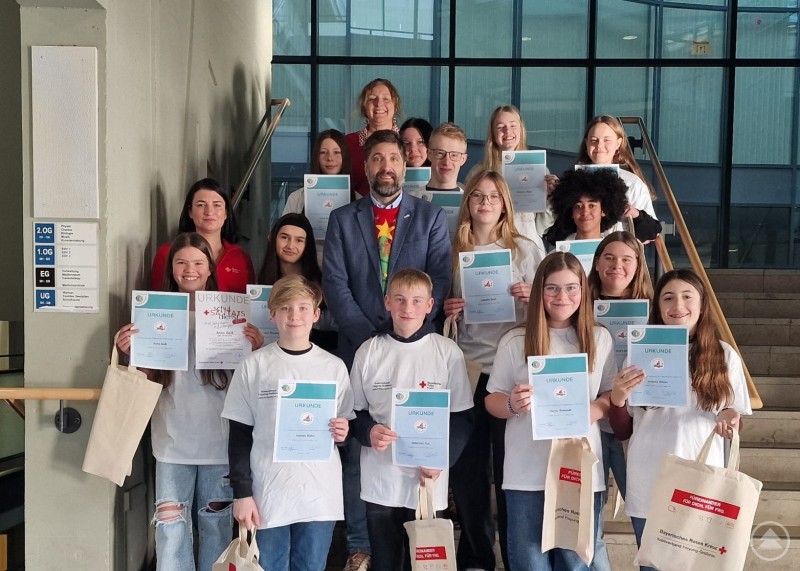 Stolz präsentieren die Grafenauer Realschülerinnen und Realschüler zusammen mit BRK-Ausbilderin Eva Feicht, den verantwortlichen Lehrkräfte Angela Fruth-Würzinger und Birgit Gsödl (nicht im Bild) sowie Schulleiter Ferdinand Klingelhöfer (von li.) ihre Urkunde zur Ernennung als neue Schulsanitäter. Gruppe von Schülerinnen und Schülern steht auf einer Treppe im Schulgebäude und hält Urkunden in die Kamera, dahinter stehen mehrere Erwachsene. Einige tragen Tragetaschen.