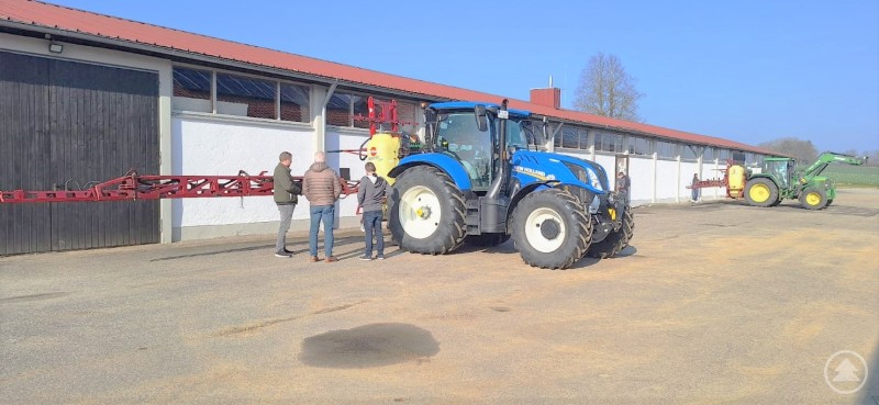 Praktische Prüfung an der Pflanzenschutzspritze in Rotthalmünster Das Bild zeigt einen blauen Traktor mit einer roten Pflanzenschutzspritze, drei Personen, die sich daneben unterhalten, und im Hintergrund einen grünen Traktor mit Spritze. Die Szene spielt sich vor einer langen Hofgebäudezeile bei sonnigem Wetter ab.