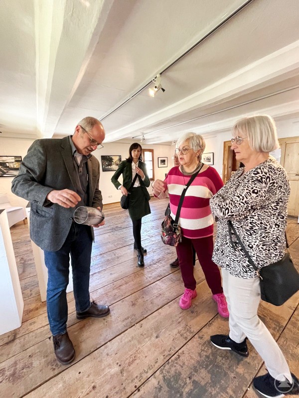 Die Besucher des Kunstmarktes konnten auch die aktuelle Ausstellung Intentionen bestaunen Das Bild zeigt eine kleine Besuchergruppe in einer Ausstellung, die offenbar im Rahmen eines Kunstmarkts stattfindet. Ein Mann im Jackett, vermutlich der Künstler oder ein Museumsführer, erklärt den interessierten Besucherinnen ein Ausstellungsstück – ein Glasobjekt, das er in der Hand hält. Die Umgebung mit Holzdielenboden, dezentem Licht und gerahmten Bildern an den Wänden vermittelt eine ruhige, konzentrierte Atmosphäre. Die Besucherinnen hören aufmerksam zu und wirken engagiert. Im Hintergrund betrachtet eine weitere Besucherin die Kunstwerke. Die Szene spiegelt Wissensvermittlung, kulturelles Interesse und die persönliche Nähe zwischen Kunst und Publikum wider.