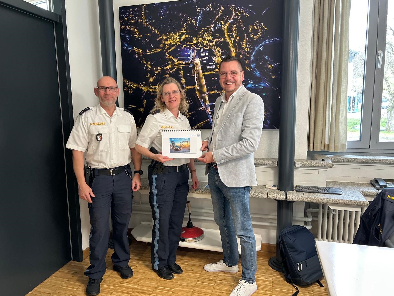 PHK Georg Binder und PORin Ingrid Grötzinger übergeben Waldkirchens Bürgermeister Heinz Pollak den aktuellen Sicherheitsbericht im Rathaus. Zwei Polizeibeamte und ein Mann in Zivilkleidung stehen in einem Besprechungsraum vor einem großen Wandbild. Der Mann in der Mitte hält ein Dokument in den Händen. Alle drei lächeln in die Kamera.