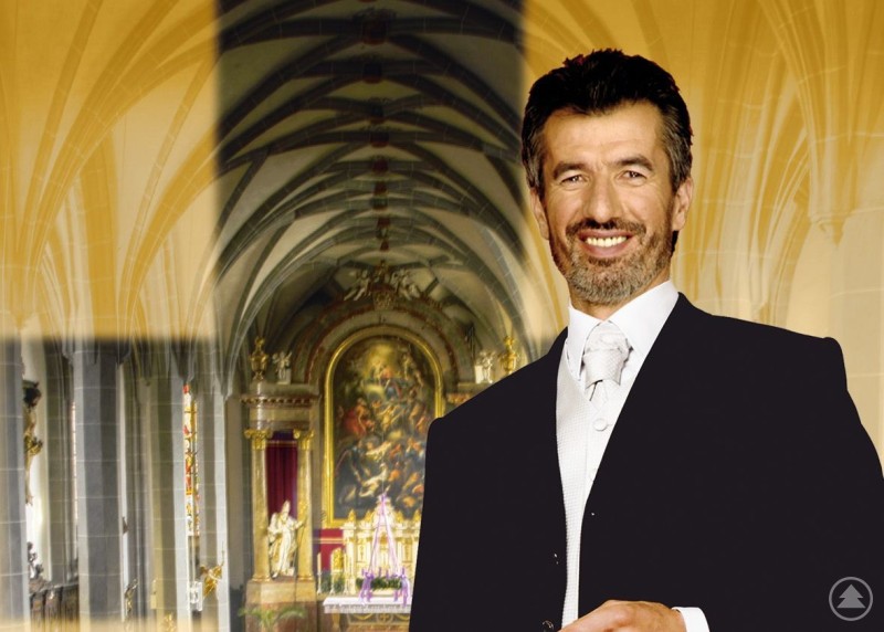 Ein Mann im dunklen Anzug mit silberner Krawatte lächelt in die Kamera. Im Hintergrund ist das Innere einer reich verzierten Kirche mit Altar und Gemälden zu sehen.