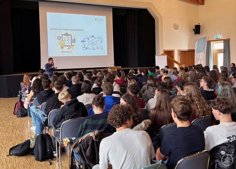 Prof. Dr. Markus Lemberger vermittelte auf kurzweilige und schülernahe Weise komplexe Inhalte des Projektmanagements. Viele Schülerinnen und Schüler sitzen in einem großen Saal und hören aufmerksam einem Vortrag zu. Auf der Leinwand ist eine Präsentation mit Illustrationen zum Thema Projektmanagement zu sehen.