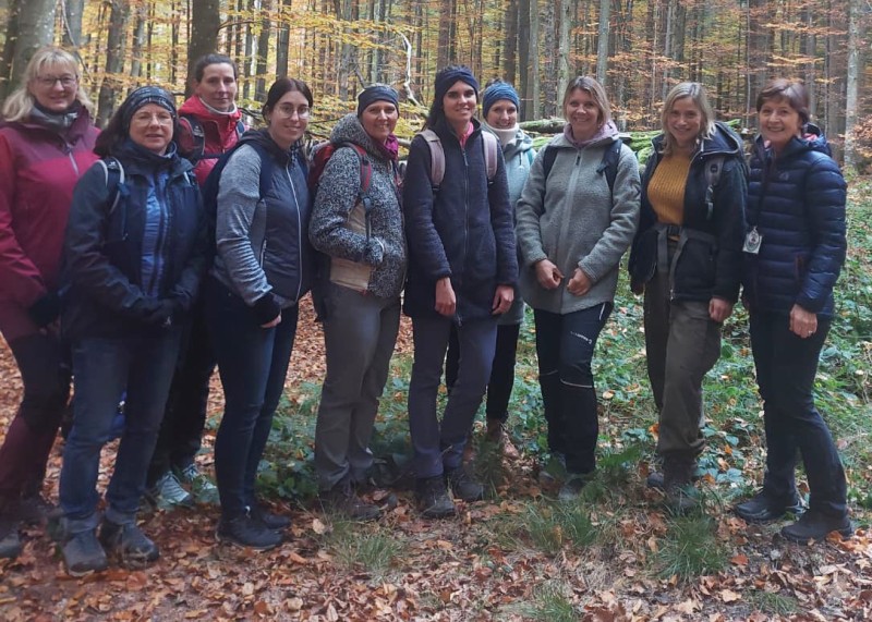 Einblicke in das Reich der Pilze erhielten die Teilnehmerinnen bei der exklusiven Fortbildung für Partner-Schulen und -Kindergärten. Eine Gruppe von zehn Frauen steht im herbstlichen Wald. Sie tragen wetterfeste Kleidung und Rucksäcke, lächeln in die Kamera und stehen auf einem mit buntem Laub bedeckten Waldboden. Im Hintergrund sind hohe, lichtdurchflutete Bäume zu sehen.
