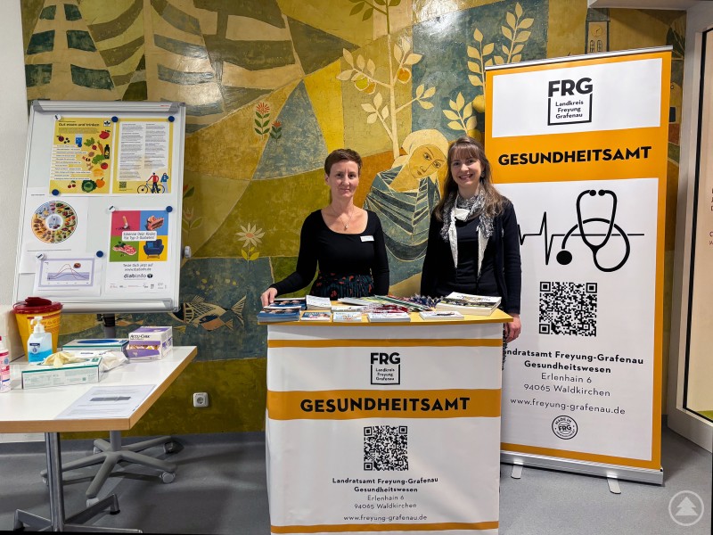 v.l.: Christine Hackinger und Dr. med. Karin Drachsler gaben am Weltdiabetestag praxisnahe Hinweise rund um Ernährung und Diabetesprävention. Zwei Mitarbeiterinnen des Gesundheitsamts Freyung-Grafenau stehen an einem Infostand zum Weltdiabetestag. Auf dem Tisch liegen Infomaterialien, im Hintergrund ein farbiges Wandmosaik und Informationsplakate.