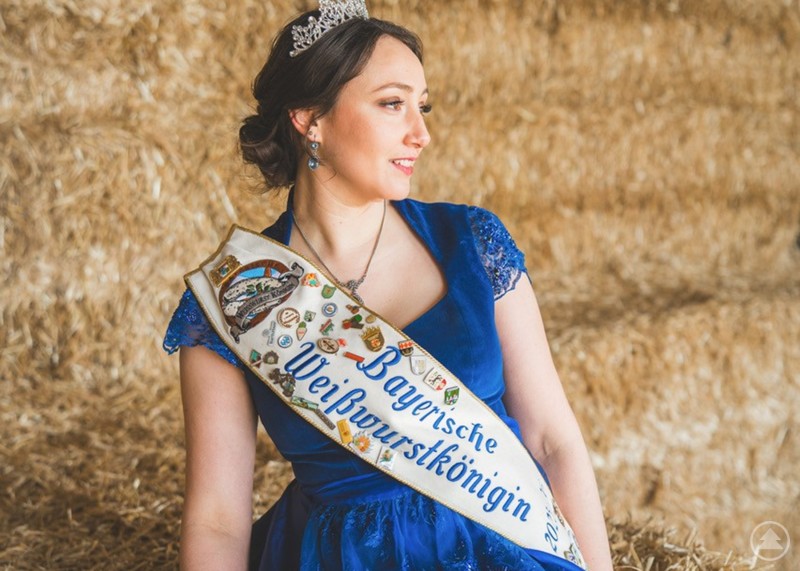 Die Bayerische Weißwurstkönigin wirbt für die Bewerbung und die Krönung in Bodenmais. Eine Frau im blauen Dirndl mit Krone und Schärpe Bayerische Weißwurstkönigin sitzt vor gestapeltem Heu und blickt zur Seite.