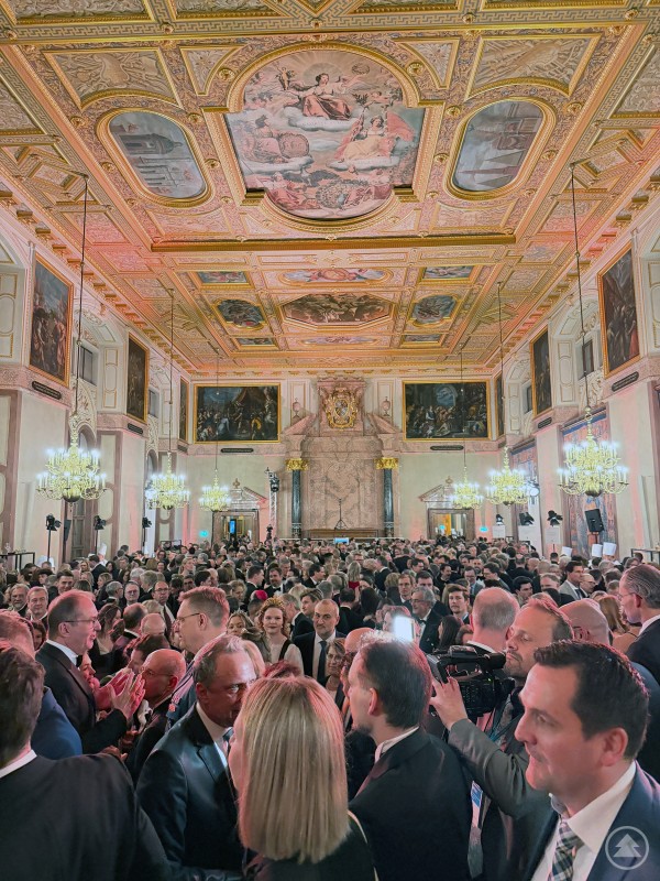 Ein Blick in den vollbesetzten Kaisersaal der Residenz in München Blick in einen großen, festlich beleuchteten Saal mit vergoldeter Decke und zahlreichen Gemälden an den Wänden. Viele elegant gekleidete Gäste stehen dicht gedrängt im Raum.