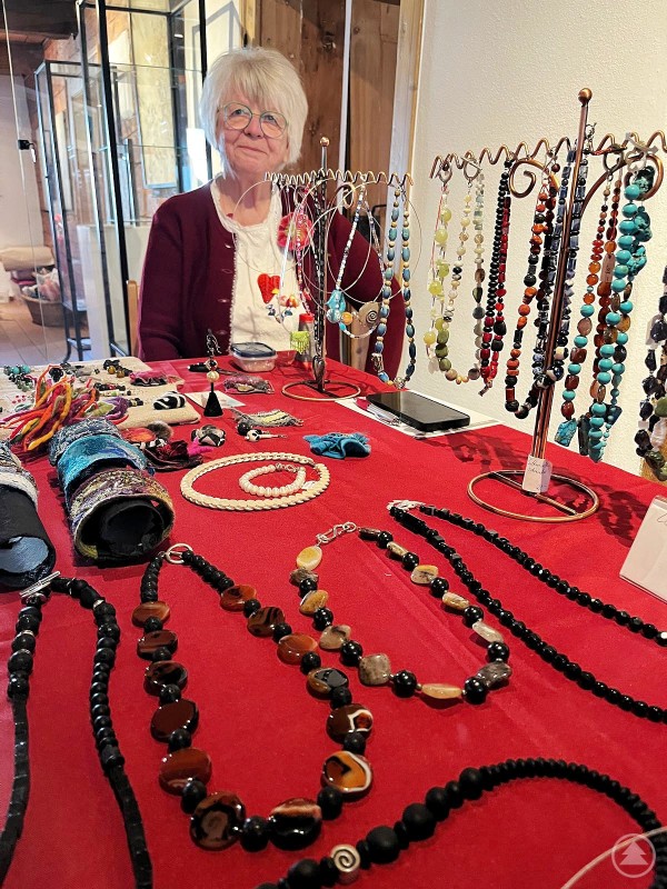 Zahlreiche Künstler und Kunsthandwerker boten ihre Werke zum Verkauf an Das Bild zeigt eine ältere Dame an einem kunstvoll gestalteten Verkaufsstand, der im Rahmen eines Kunst- oder Handwerkermarkts entstanden ist. Auf einem roten Tuch präsentiert sie eine große Auswahl an selbst gefertigten Schmuckstücken – darunter Ketten, Armbänder und Anhänger aus Perlen, Halbedelsteinen und Draht. Die Schmuckstücke sind farbenfroh, kreativ und mit Liebe zum Detail gefertigt. Die Verkäuferin wirkt freundlich und stolz auf ihre Arbeiten. Im Hintergrund sind Vitrinen und ein rustikales Ambiente erkennbar, was auf eine Ausstellung oder einen historischen Veranstaltungsort hinweist. Die Szene strahlt handwerkliches Können, Persönlichkeit und Freude am kreativen Schaffen aus.