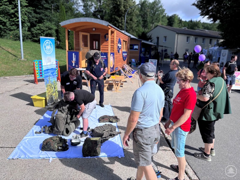 Die gesamte persönliche Ausrüstung eines Soldaten musste in den Rucksack – und zwar so, dass nichts mehr klappert Besucher beobachten eine Station, bei der ein Kind militärisches Gepäck in einen Rucksack packt; im Hintergrund steht ein Holzhaus mit TonYversum-Branding.