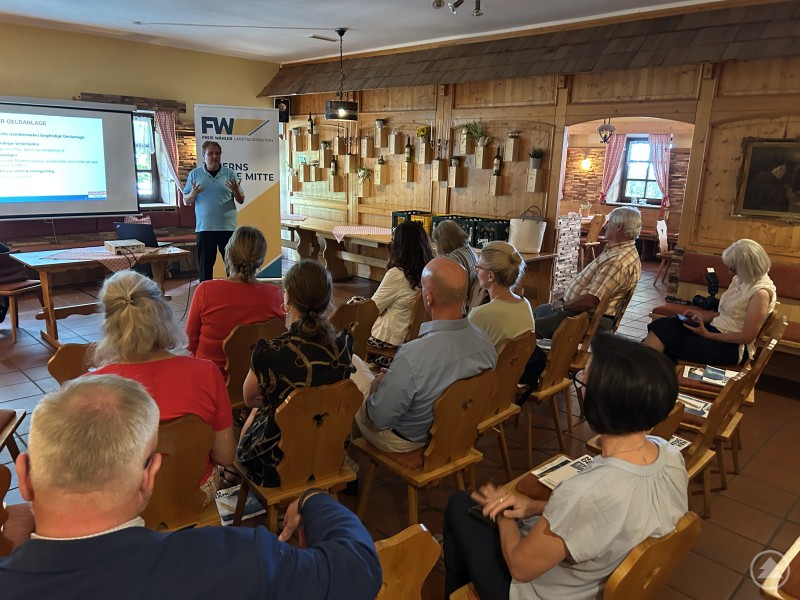 Sascha Straub referiert im Deggendorfer Krahwirt über geschlechtergerechte Altersvorsorge – eingeladen von der FW-Landtagsfraktion. Ein Mann hält in einem Gastraum vor einem Plakat der FW-Landtagsfraktion einen Vortrag über Geldanlage, das Publikum sitzt auf Holzstühlen.