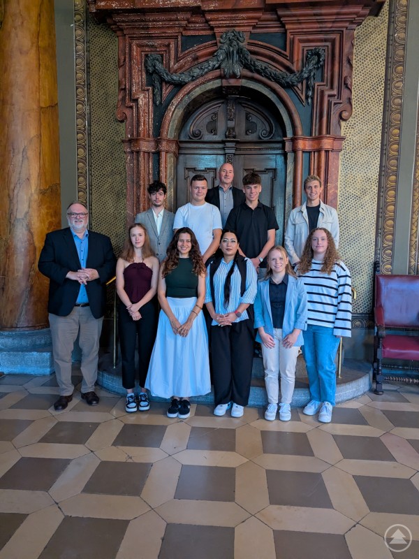 Oberbürgermeister Jürgen Dupper (links) begrüßt zusammen mit Personalreferent Herbert Zillinger (hinten) und Ausbildungsleiter Maximilian Drechsler (rechts, zweite Reihe) die neuen Anwärterinnen, Anwärter und Auszubildenden der Stadt Passau. Eine Gruppe von zwölf Personen, darunter junge Erwachsene und drei ältere Männer, posiert für ein Gruppenfoto vor einer verzierten Holztür im historischen Ambiente.