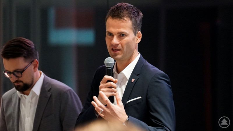 Das zweite Bild zeigt zwei Männer im Anzug. Der eine im Vordergrund hält ein Mikrofon und spricht, während er mit der anderen Hand gestikuliert. Der zweite Mann im Hintergrund schaut nach unten. Auch dieses Bild vermittelt den Eindruck einer offiziellen Veranstaltung oder eines Kongresses.