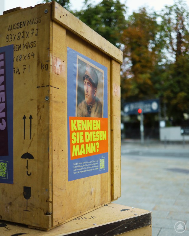 Mit der Frage „Kennen Sie diesen Mann?“ bittet das Oberhausmuseum um Hinweise zu einem unbekannten Porträt. Nahaufnahme einer Holztransportkiste mit einem Plakat „Kennen Sie diesen Mann?“ und dem Porträt eines Soldaten.