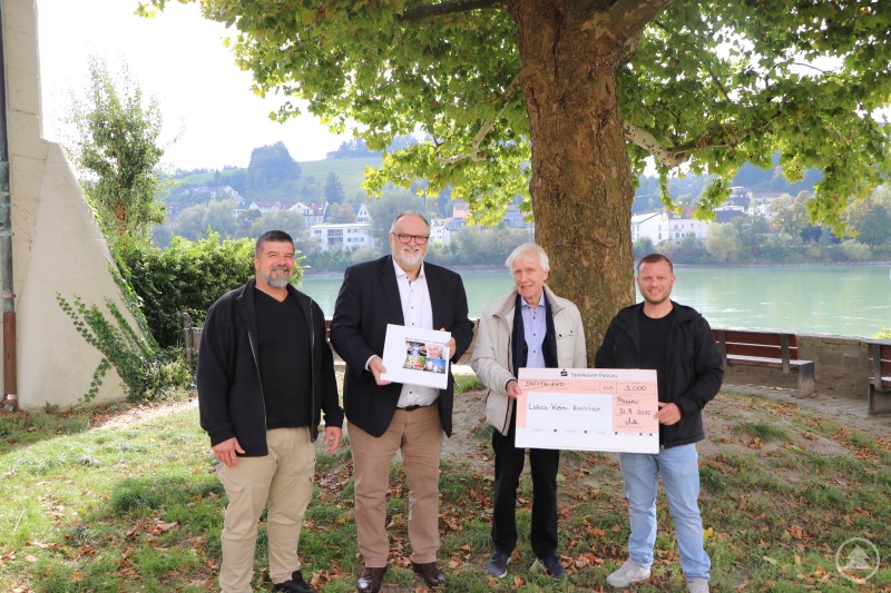 Bei der Scheckübergabe: Oberbürgermeister Jürgen Dupper (3. v. r.) nimmt zusammen mit dem Leiter des Lukas-Kern-Kinderheims, Andreas Effenberg (rechts), und seinem Stellvertreter, Jürgen Silzer (links), den symbolischen Scheck von Gisbert Burgstaller entgegen. Vier Männer stehen im Freien unter einem großen Baum am Donauufer. Einer von ihnen hält einen symbolischen Scheck, ein anderer ein Foto. Im Hintergrund sind Fluss, Ufer und die Stadt Passau zu sehen.