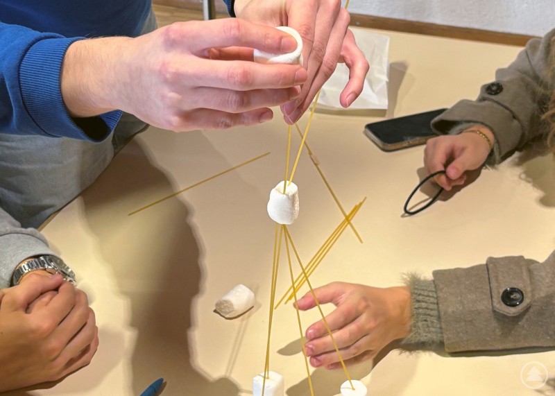 Die Schülerinnen und Schüler erhielten die Aufgabe, aus Spaghetti und Marshmallows einen möglichst hohen Turm zu konstruieren. Mehrere Personen bauen gemeinsam mit Spaghetti und Marshmallows einen Turm. Nur die Hände der Teilnehmenden sind zu sehen, wie sie konzentriert am Projekt arbeiten.
