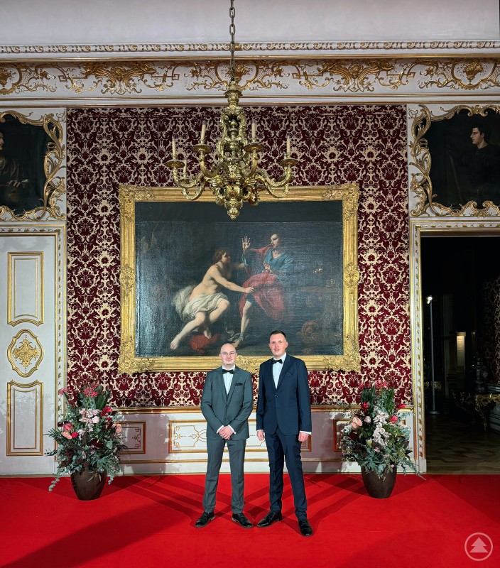 Zwei M&auml;nner im Smoking stehen auf einem roten Teppich in einem prunkvollen Saal. Hinter ihnen h&auml;ngt ein gro&szlig;es Gem&auml;lde in Goldrahmen, seitlich sind Blumenarrangements platziert.