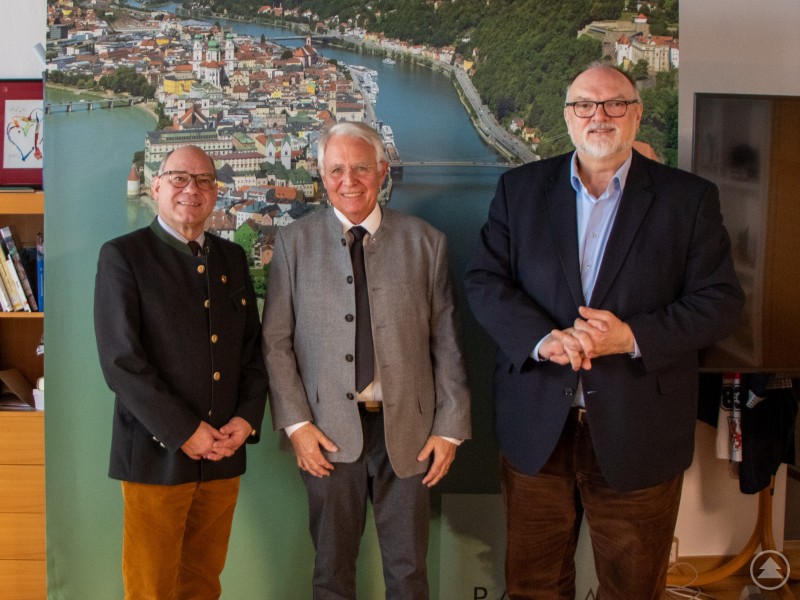 Oberbürgermeister Jürgen Dupper empfängt (v. l.) stellv. Bruderschaftsmeister Ralf Schützenberger und Bruderschaftsmeister Dr. Ludwig Bauer im Amtszimmer. Drei Männer stehen nebeneinander im Amtszimmer des Rathauses vor einer großformatigen Stadtansicht von Passau. Sie tragen Anzug beziehungsweise Trachtenjanker und lächeln in die Kamera.