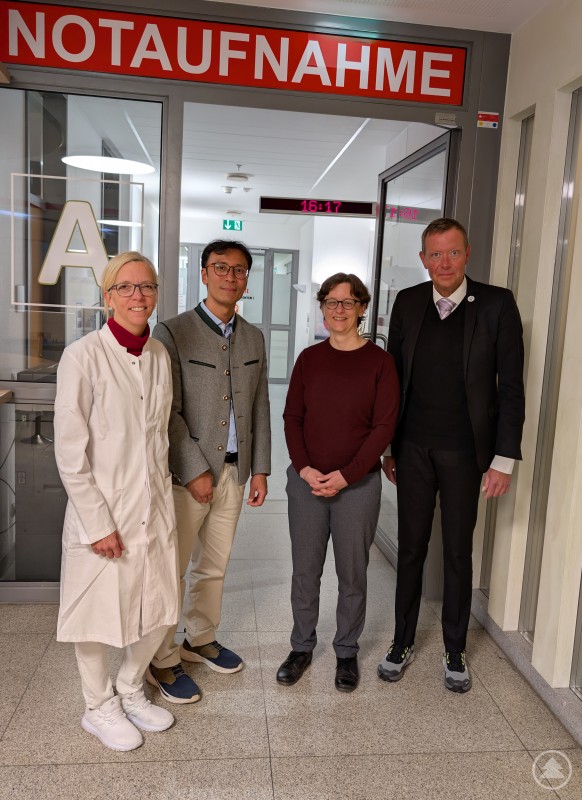 v.l.: Dr. med. Simone Götz, Prof. Dr. Thiha Aung, Prof. Dr. Katja Goldflam, Marcus Plaschke (Fehlt: Dr. med. Andrea Pfeiffer, da verhindert wg. eines akuten Notfalls) Vier Personen stehen im Eingangsbereich der Notaufnahme des Klinikums Freyung. Hinter ihnen ist das rote Schild Notaufnahme zu sehen. Die Gruppe besteht aus einer Ärztin im weißen Kittel sowie drei weiteren Personen aus Medizin und Klinikleitung, die sich zu einem Besuch und fachlichen Austausch vor Ort getroffen haben.