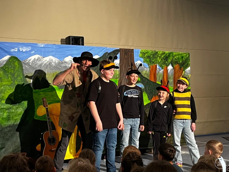 Theatermacher Toni Tanner mit den Insektenkindern (v. l. Korbinian Wippich, David Hilpert, Mia Joachimsthaler und Nina Janka) Ein Musiker steht mit mehreren Kindern auf einer kleinen Bühne vor einer bemalten Kulisse mit Bergen und Wald. Die Kinder tragen Kostüme und spielen Insektenrollen, während im Vordergrund weitere Kinder als Publikum sitzen.
