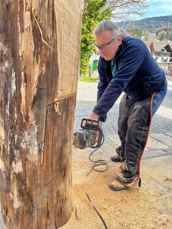 Holzkünstler Walter Schreiner in Aktion Das Bild zeigt den Holzkünstler Walter Schreiner bei der Arbeit an einem massiven Baumstamm im Freien. Mit Schutzbrille, Gehörschutz und Motorsäge ausgestattet, konzentriert er sich auf einen präzisen Schnitt, während Sägemehl zu seinen Füßen liegt. Die Szene spielt sich auf einem gepflasterten Platz inmitten einer ländlichen Kulisse mit Hügeln, Wäldern und Häusern im Hintergrund ab. Das Bild vermittelt handwerkliches Können, kreative Energie und die Nähe zur Natur – ein authentischer Moment der Holzbildhauerei in Aktion.