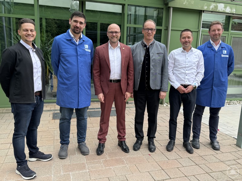 Minister Dr. Fabian Mehring (Mitte) und Abgeordneter Martin Behringer (rechts daneben) beim Besuch des LINHARDT-Stammwerks in Viechtach mit Unternehmensvertretern. Sechs Männer stehen nebeneinander vor einem modernen, grünen Firmengebäude. Zwei von ihnen tragen blaue Arbeitskittel mit LINHARDT-Logo, die anderen sind in Businesskleidung oder im Trachtenjanker gekleidet. Alle blicken freundlich in die Kamera.