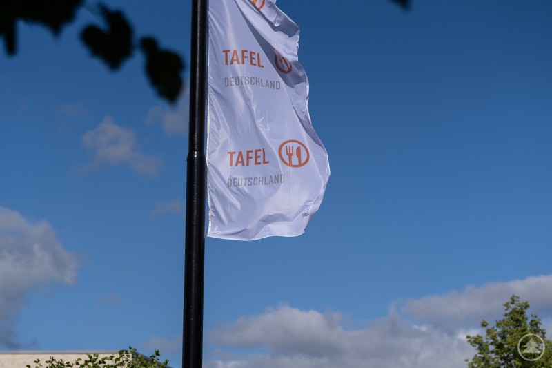 Weiße Fahne mit dem Logo und Schriftzug "TAFEL DEUTSCHLAND" weht im Wind vor blauem Himmel, teilweise von Baumzweigen umrahmt. Weiße Fahne mit dem Logo und Schriftzug "TAFEL DEUTSCHLAND" weht im Wind vor blauem Himmel, teilweise von Baumzweigen umrahmt.